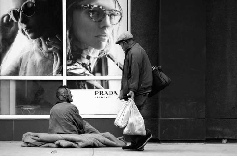 Man with shopping bags walking past a beggar on the sidewalk — UPSC Mains essay topic Need Brings Greed, and If Greed Increases It Spoils Breed.