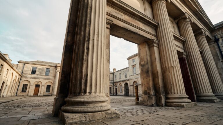 A monumental stone portico with heavy columns seen from below, framing an empty administrative courtyard beyond.