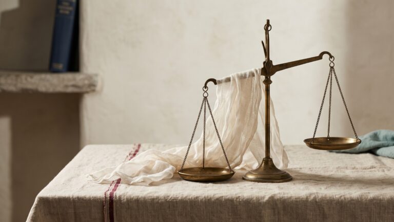 A brass balance scale on a linen-covered table with one pan hanging slightly higher, a length of translucent gauze draped loosely over the fulcrum.