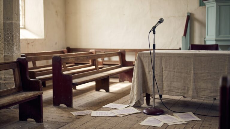 A small-town meeting hall with rows of wooden benches, a single microphone on a cloth-covered table, and loose cyclostyled sheets scattered on the floor.