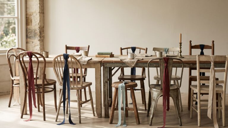 A long table assembled from mismatched sections with chairs of many different styles drawn up to it, coloured ribbons knotted at one end.