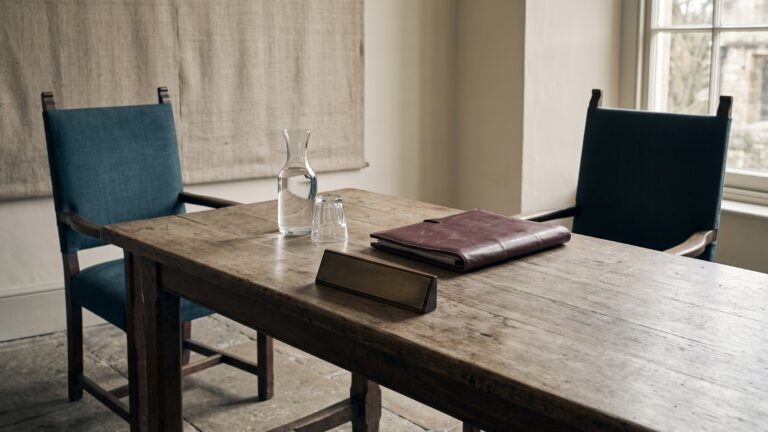 A diplomatic meeting table with a leather document folder, a blank nameplate, a water carafe and two chairs facing each other.