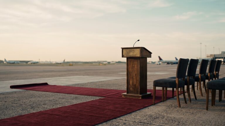 An airport tarmac at dawn with a red carpet unrolled beside a bare lectern and a waiting line of empty chairs.
