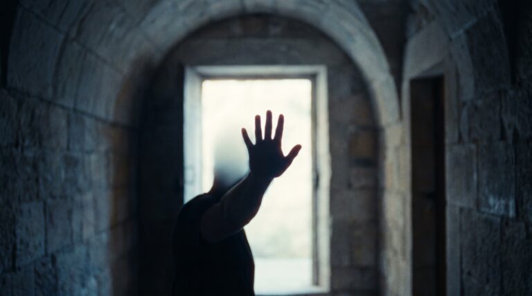 A raised open palm silhouetted against a bright doorway in a dim corridor