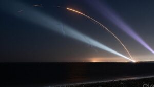 Two missile trails over dark coastal water at night, crossed by air-defence searchlights, with a faint city glow on the horizon