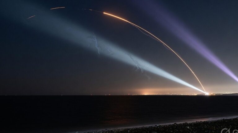 Two missile trails over dark coastal water at night, crossed by air-defence searchlights, with a faint city glow on the horizon