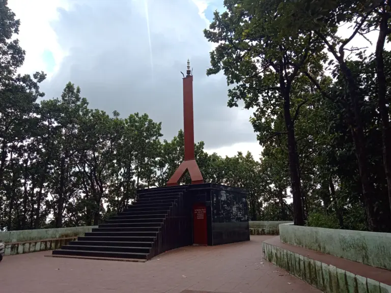 A tall red obelisk rises behind a dark stairway and memorial panels at the Khalanga War Memorial, framed by tall trees.