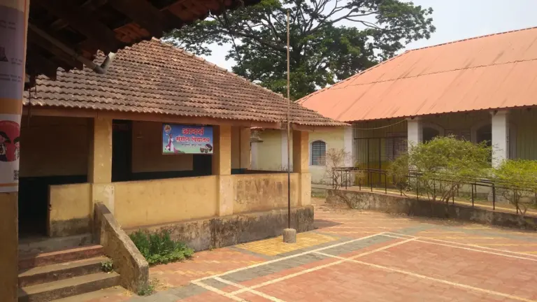 Tiled-roof village school buildings with ochre walls and a covered veranda face a paved courtyard, with a large tree behind.