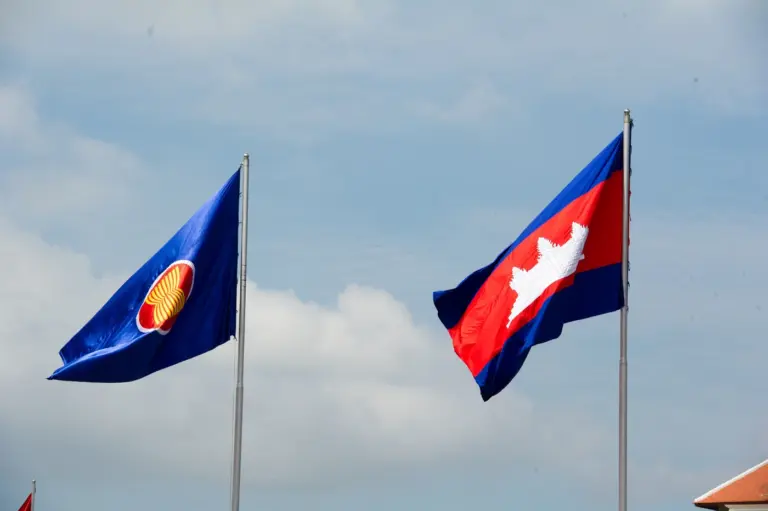 The blue ASEAN flag and the red and blue flag of Cambodia fly on tall poles against a cloudy sky.