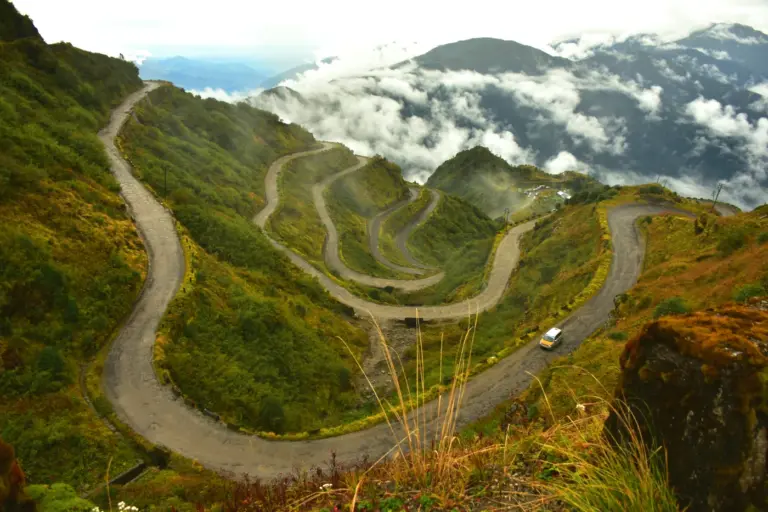 Hairpin loops of a mountain road zigzagging down green Himalayan slopes in Sikkim, with a car on a lower bend
