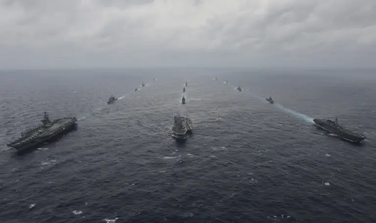 Flat-decked carriers and a long line of warships sail in formation across a gray sea under an overcast sky.