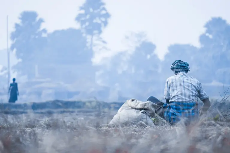 A farmer in a checked shirt and head cloth sits beside a sack in a hazy, dry field, seen from behind.