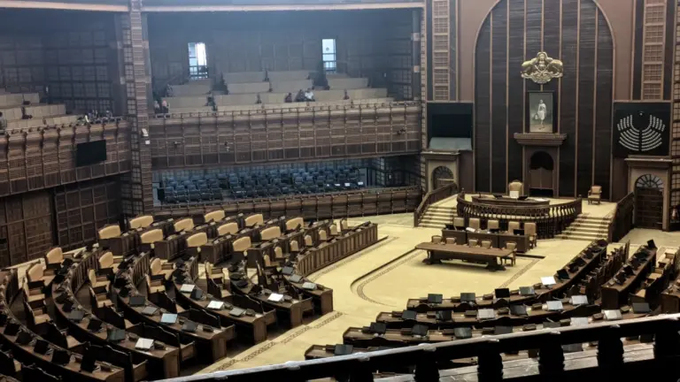 The empty Kerala Legislative Assembly chamber, with curved rows of wooden desks and chairs facing the raised Speaker's dais and wood-paneled walls.