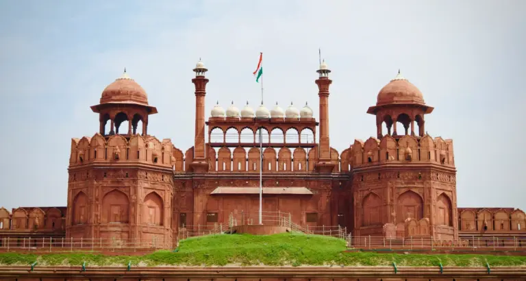 The red sandstone Lahori Gate of the Red Fort in Delhi, with twin domed towers and an Indian flag above a green lawn.