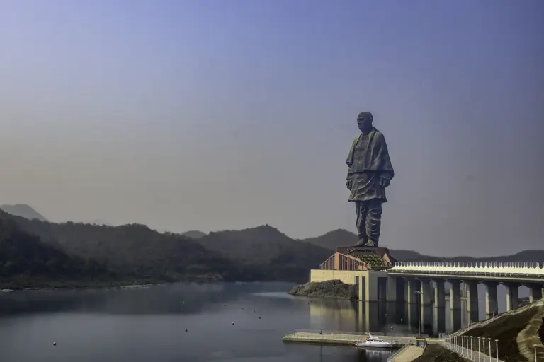 The Statue of Unity rises above calm river water in Gujarat, with hazy hills behind and a long walkway leading to its base.