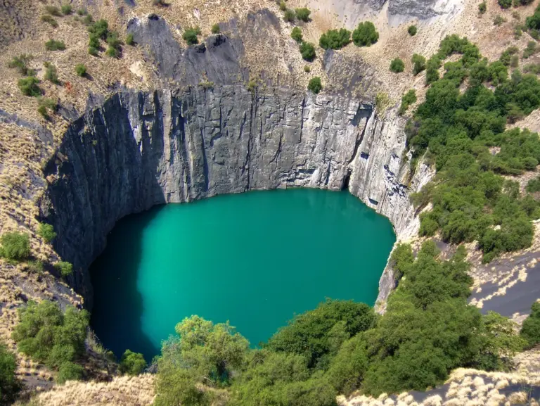 The flooded Big Hole open-pit mine in Kimberley, South Africa, with sheer rock walls and turquoise water ringed by green bushes.