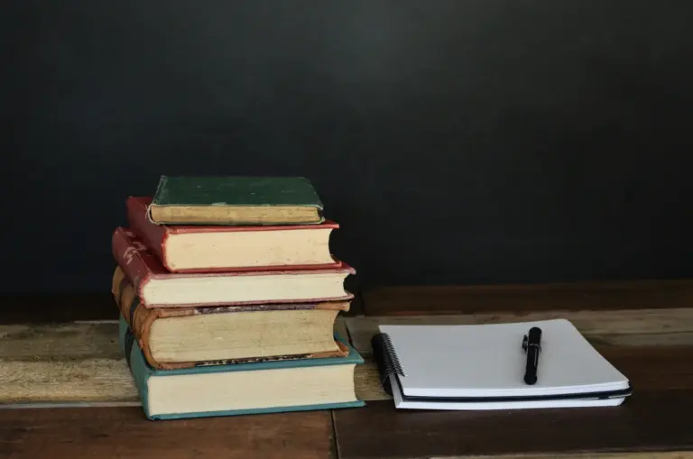Pile of books beside white printer paper and a black ballpoint pen. Photo by Debby Hudson on Unsplash via Unsplash.