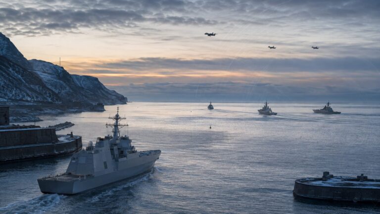 A new gray destroyer leaving an east-coast harbor while distant ships and aircraft conduct a coordinated multidomain exercise