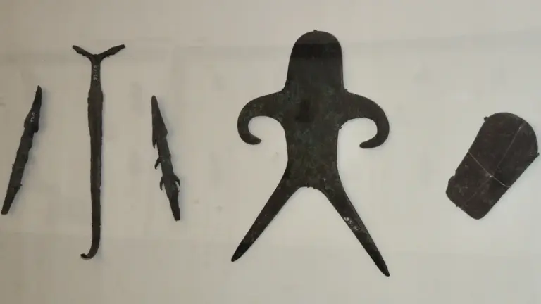 Ancient tools, including two barbed points, a forked rod, an anthropomorphic figure and flat axe blades, laid out on a pale museum display.