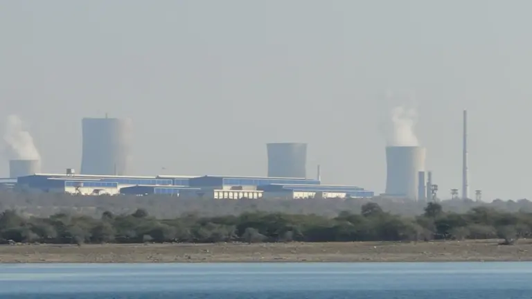 Cooling towers, a tall stack and plant buildings of the Rawatbhata nuclear power station rise beyond calm water on a hazy day.