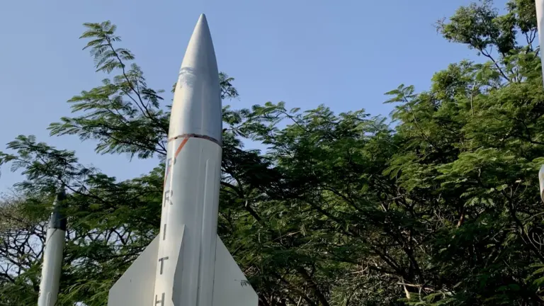 A white Prithvi missile with orange stripe markings stands upright on outdoor display, framed by leafy trees and a clear blue sky.