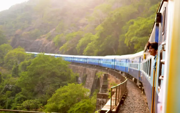 A blue Indian Railways passenger train curving over a stone viaduct through forested hills in Goa