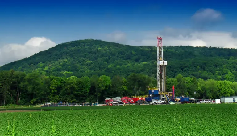 A tall drilling rig rises above trucks and equipment at the edge of a green crop field, with a wooded hill behind.