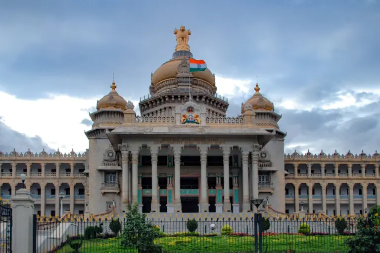 The granite facade of Vidhana Soudha in Bengaluru, with a central dome, columned portico and an Indian flag against a cloudy sky.