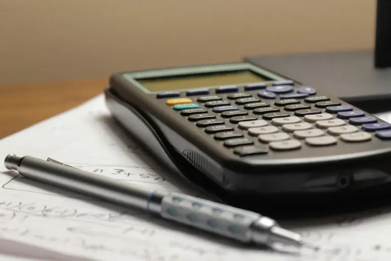 A black calculator and a silver pen rest on a sheet of handwritten figures on a wooden desk.