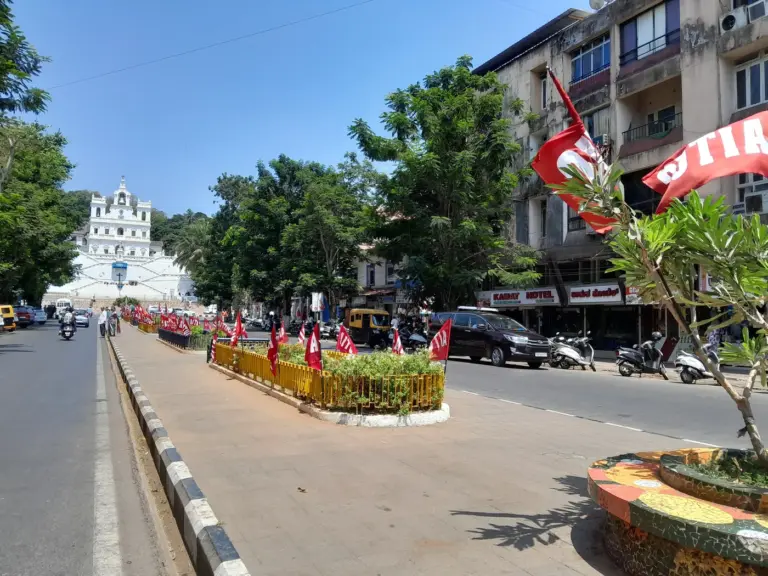 Red AITUC flags line a landscaped road divider in Panaji, Goa, with a white church facade at the end of the street.