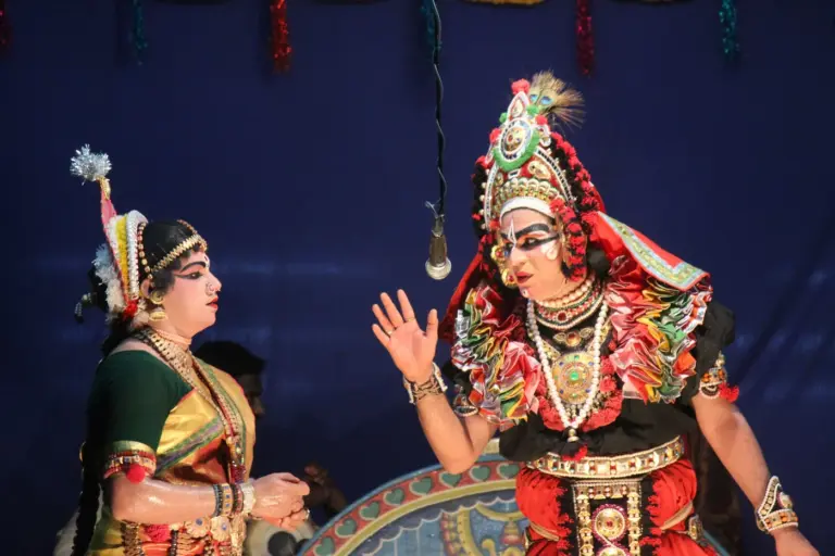 Two Yakshagana performers on stage in Udupi, Karnataka, one wearing a tall ornate crown and red-and-gold costume