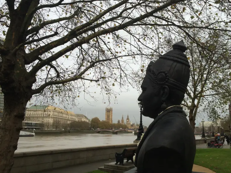 A bronze bust of Basaveshwara on the Albert Embankment in London, with the River Thames and the Houses of Parliament behind it.
