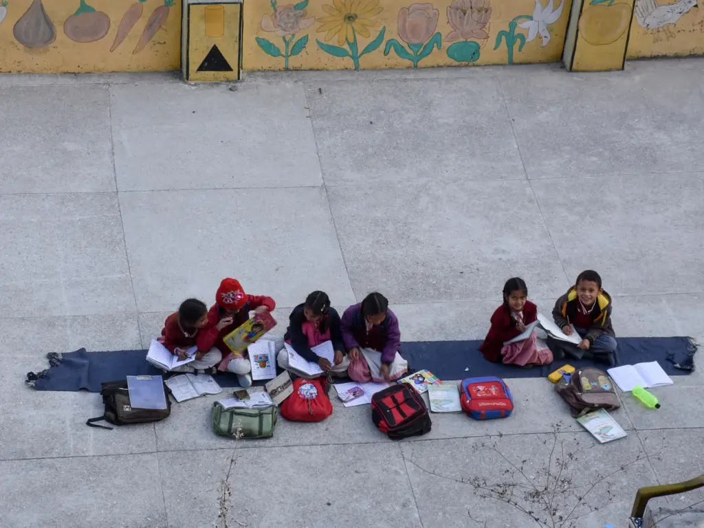 Schoolchildren sit on a mat reading their books in a sunny school courtyard in Kullu, Himachal Pradesh.